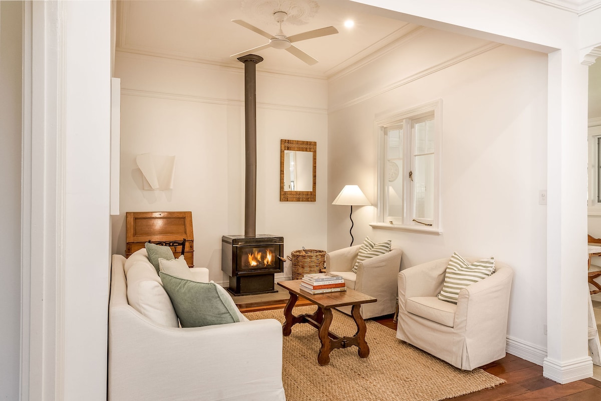 A comfortable lounge area features a cozy fireplace surrounded by a white sofa and two armchairs with decorative cushions. A wooden coffee table sits at the center, complemented by a textured area rug. Natural light enters through windows adorned with simple white curtains.