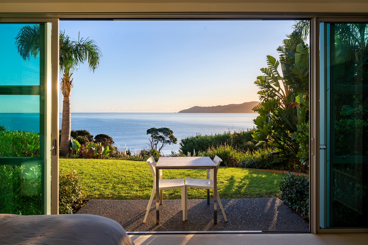 An outdoor area features a small white table and chairs set on a grassy lawn. A panoramic view of the ocean extends to the horizon, framed by lush greenery and palm trees under a clear sky, suggesting a serene and inviting atmosphere.