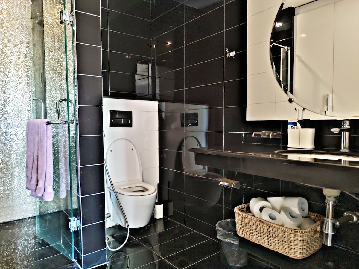 The bathroom features sleek black tiled walls, enhancing a modern aesthetic. A glass shower is visible, with textured walls adding depth. A round mirror sits above a dark countertop, with toiletries and a woven basket containing extra supplies placed below. Towels hang near the shower area.