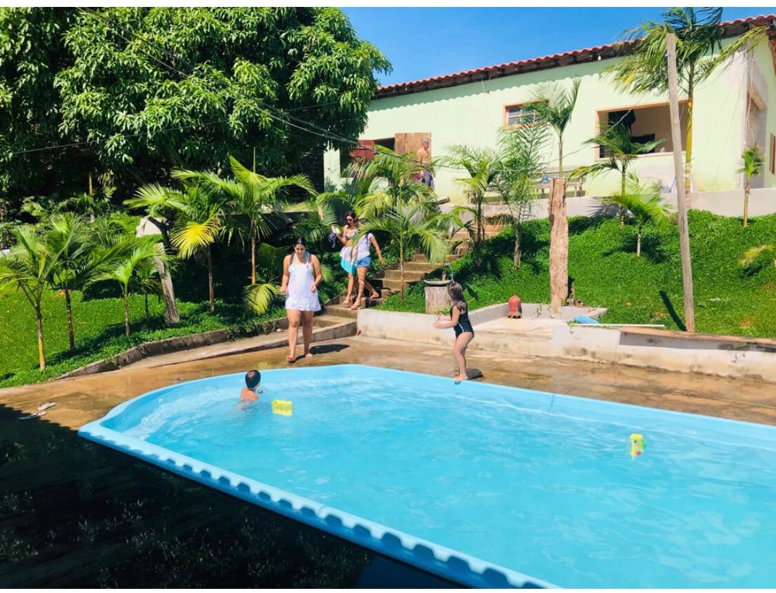 A swimming pool is framed by lush greenery, with palm trees lining the surrounding area. Two adults and a child are present near the pool, while another child swims. A home is visible in the background, surrounded by a grassy lawn.