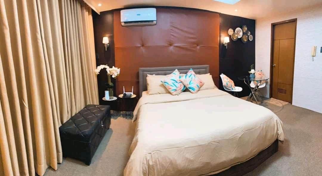 A cozy bedroom features a spacious bed with a neutral-toned duvet and decorative pillows. Soft lighting is provided by wall sconces, and an air conditioning unit is mounted above. A small accent table holds vases with flowers, while a chic ottoman is positioned at the foot of the bed.