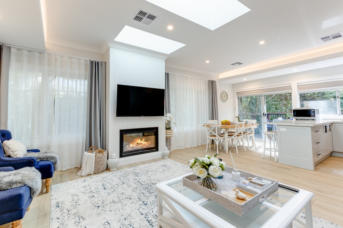 An open living space is filled with natural light from skylight windows. A modern gas fireplace is positioned beneath a wall-mounted TV. A dining area for six is visible, along with a cozy seating arrangement featuring navy chairs. Light sheer curtains soften the view of the outdoor garden.