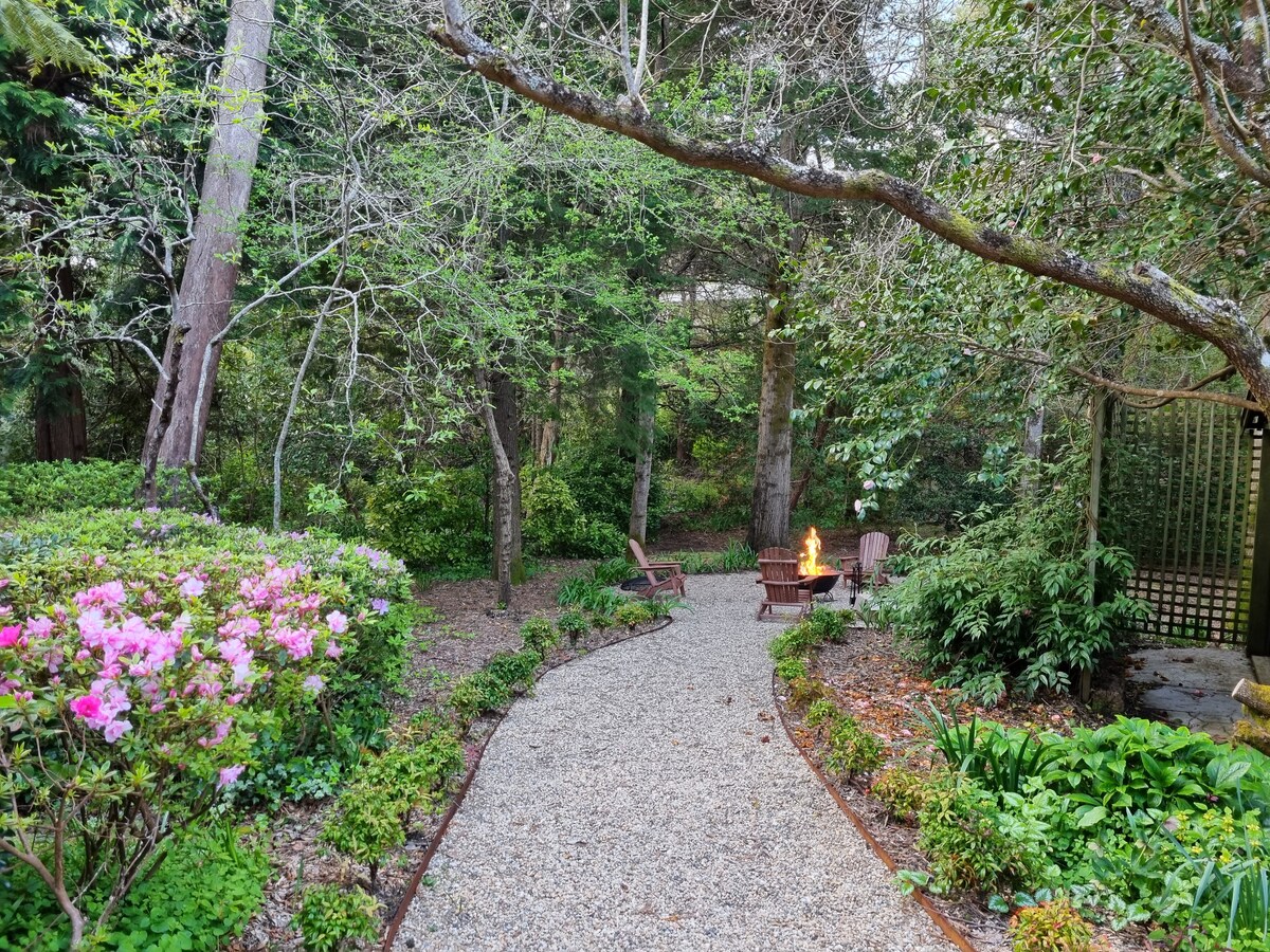 A serene garden path meanders through lush greenery, bordered by blooming flowers. Two Adirondack chairs are positioned around a circular fire pit, surrounded by trees and shrubs, creating a tranquil retreat within nature.