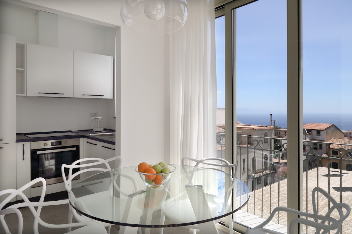 A modern dining area features a glass table surrounded by white chairs. Natural light floods the space through large windows, offering views of the sea and surrounding rooftops. A bowl of fruit adds a touch of color to the tabletop.