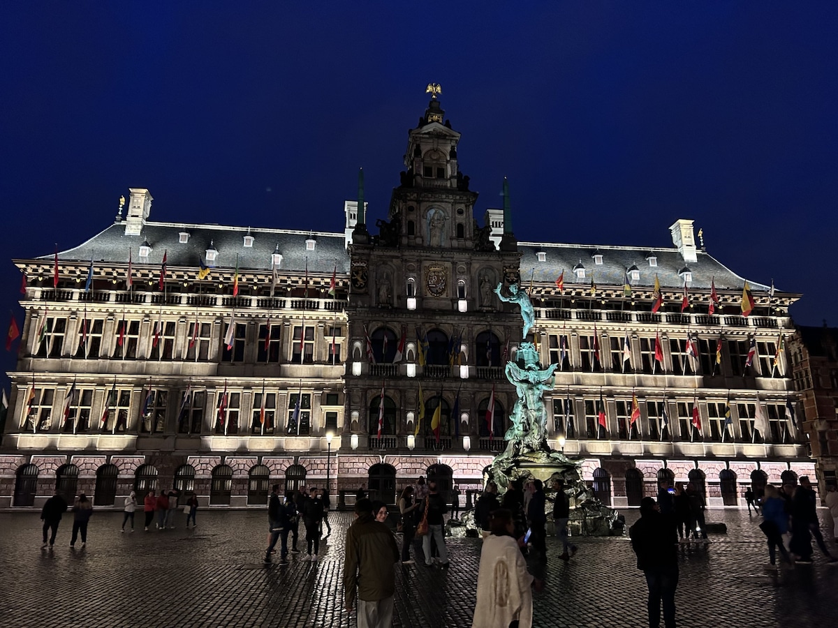 The image captures a historic building illuminated at night, showcasing intricate architectural details and decorative elements. A central fountain featuring a statue is visible in the foreground. The square is populated with visitors, adding to the lively atmosphere of the scene.