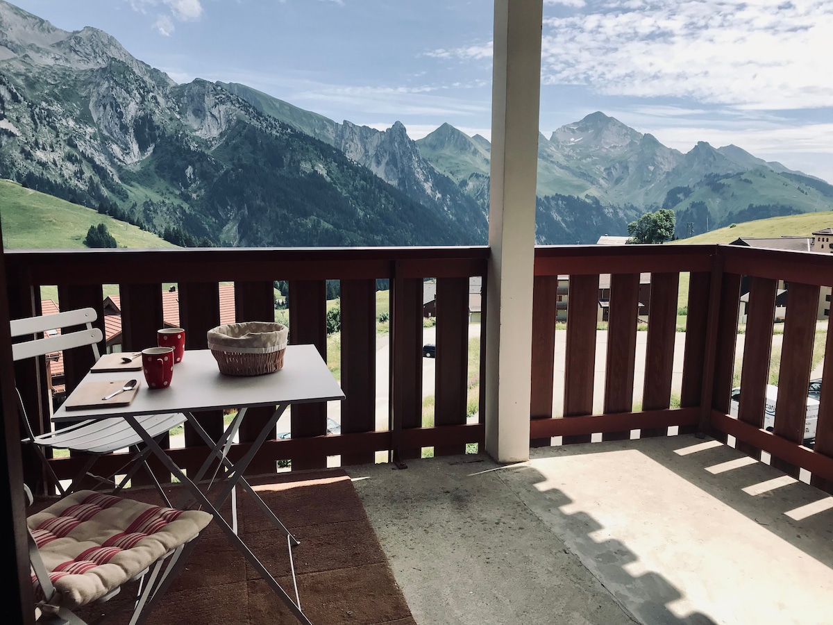 A spacious balcony is shown with a table and two chairs, offering captivating mountain views. Sunlight illuminates the area, with green hills and peaks visible in the distance, providing a serene outdoor space for relaxation.