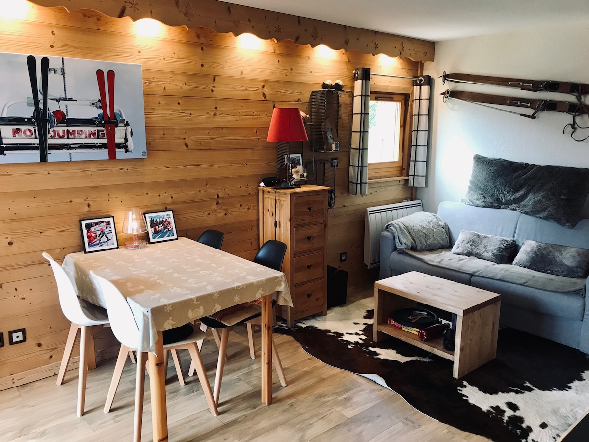 A welcoming living area features a light wooden wall adorned with vintage ski equipment. A cozy sofa sits beside a coffee table on a patterned rug. A dining table with black chairs is positioned nearby, illuminated by soft wall lighting.