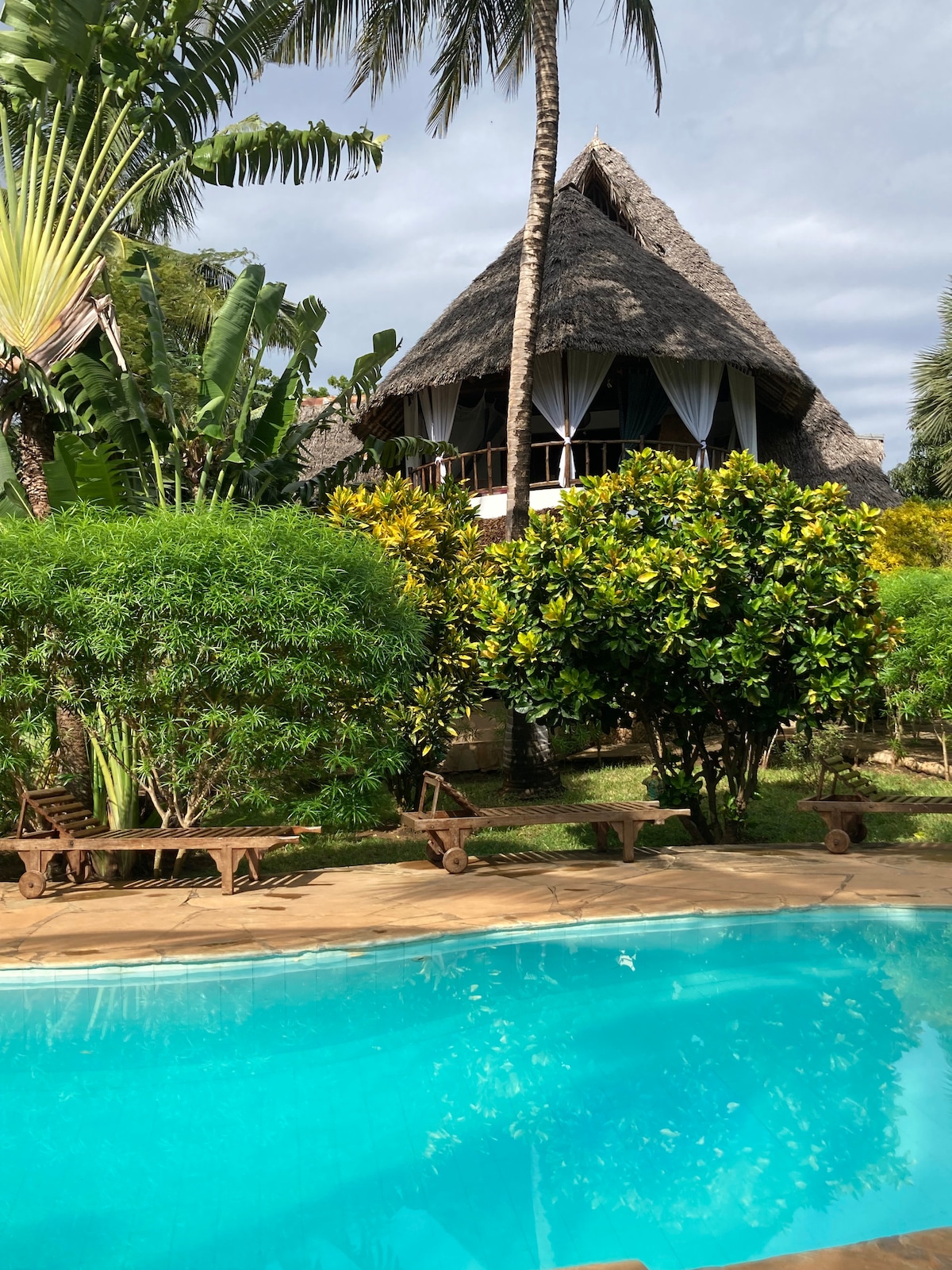 A spacious pool is surrounded by lush greenery, with a traditional thatched-roof building visible in the background. The structure features large windows and a balcony, enhancing the inviting atmosphere of the garden setting.