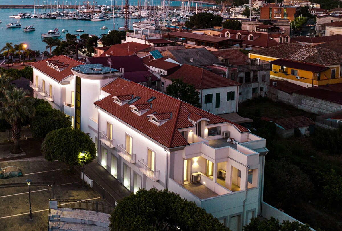 The apartment building is presented in an elegant style, featuring a white facade and terracotta roof tiles. Surrounding greenery is observed, along with a view of the marina in the background, where multiple boats are docked in the shimmering water.