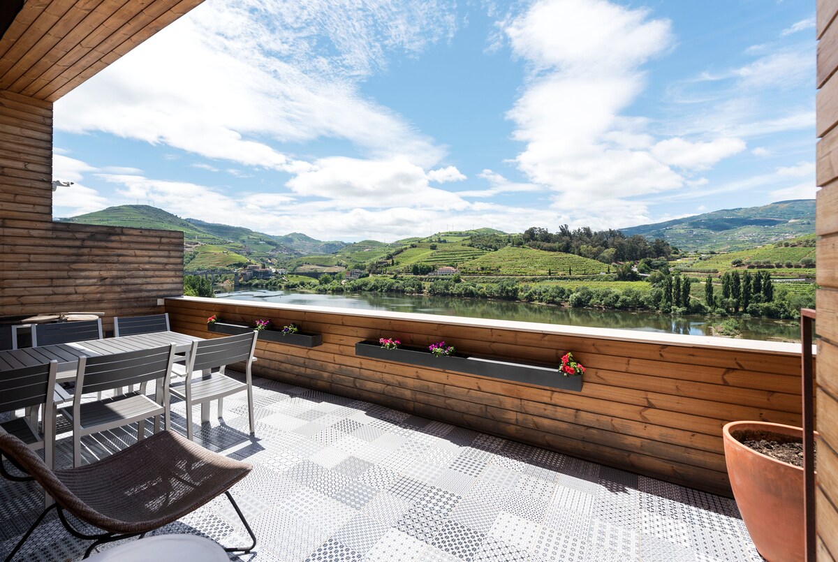 A spacious terrace overlooks serene landscapes of rolling hills and the river, with wooden paneling and tiled flooring. Outdoor seating is arranged alongside colorful flower boxes, creating a welcoming outdoor space for relaxation.
