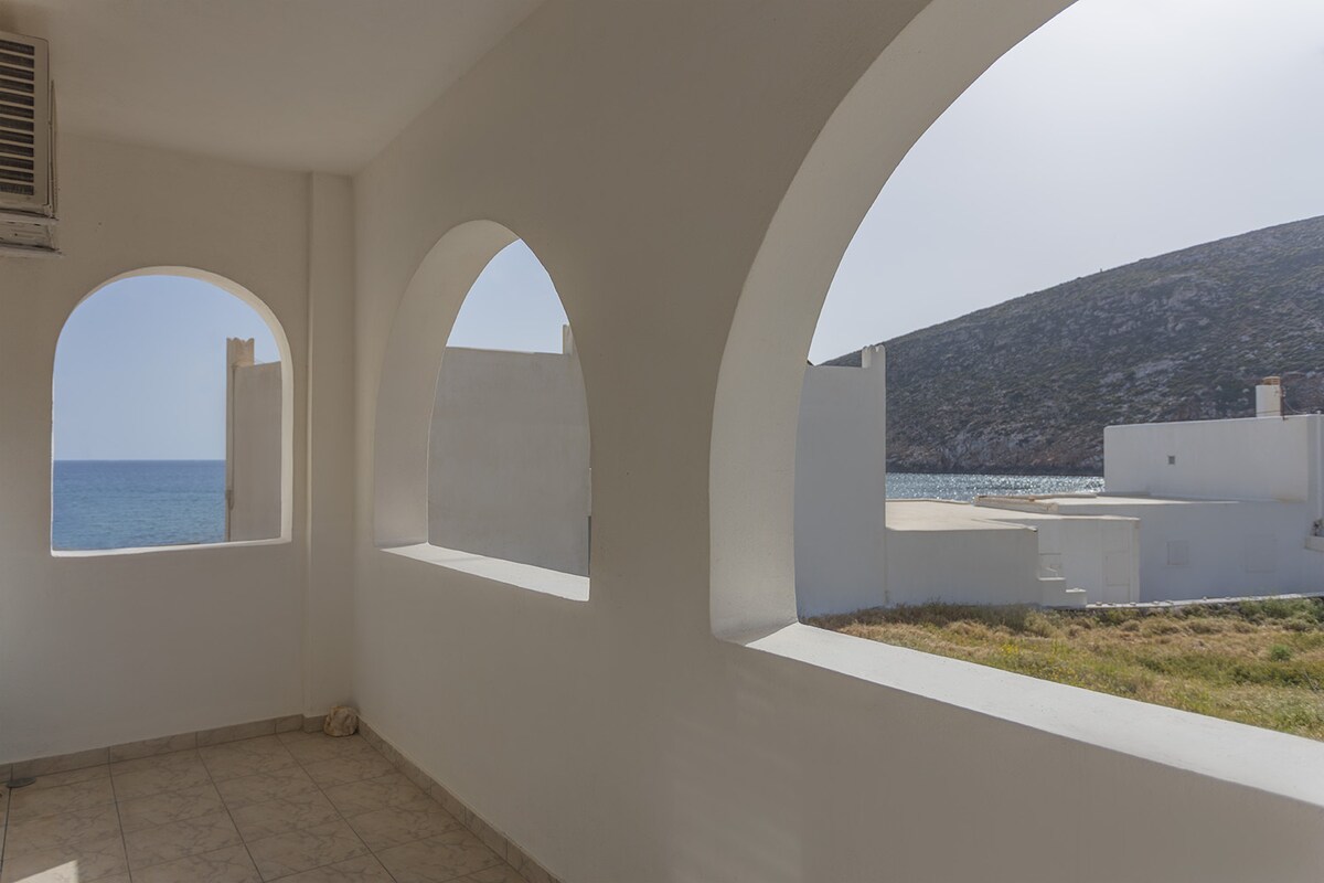 A spacious balcony features elegant arched openings, framing views of the sea and the natural landscape beyond. Soft sunlight bathes the neutral-toned tiles, creating a serene and open atmosphere. The surrounding white walls enhance the sense of openness and invite relaxation.