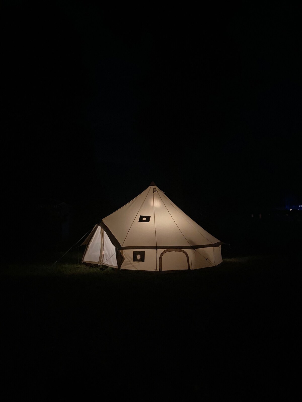 A glamping tent is softly illuminated against a dark night backdrop. The tent features a conical shape with multiple windows, allowing the glow from inside to gently spread into the surrounding area. A grassy field surrounds the tent, enhancing the serene outdoor setting.