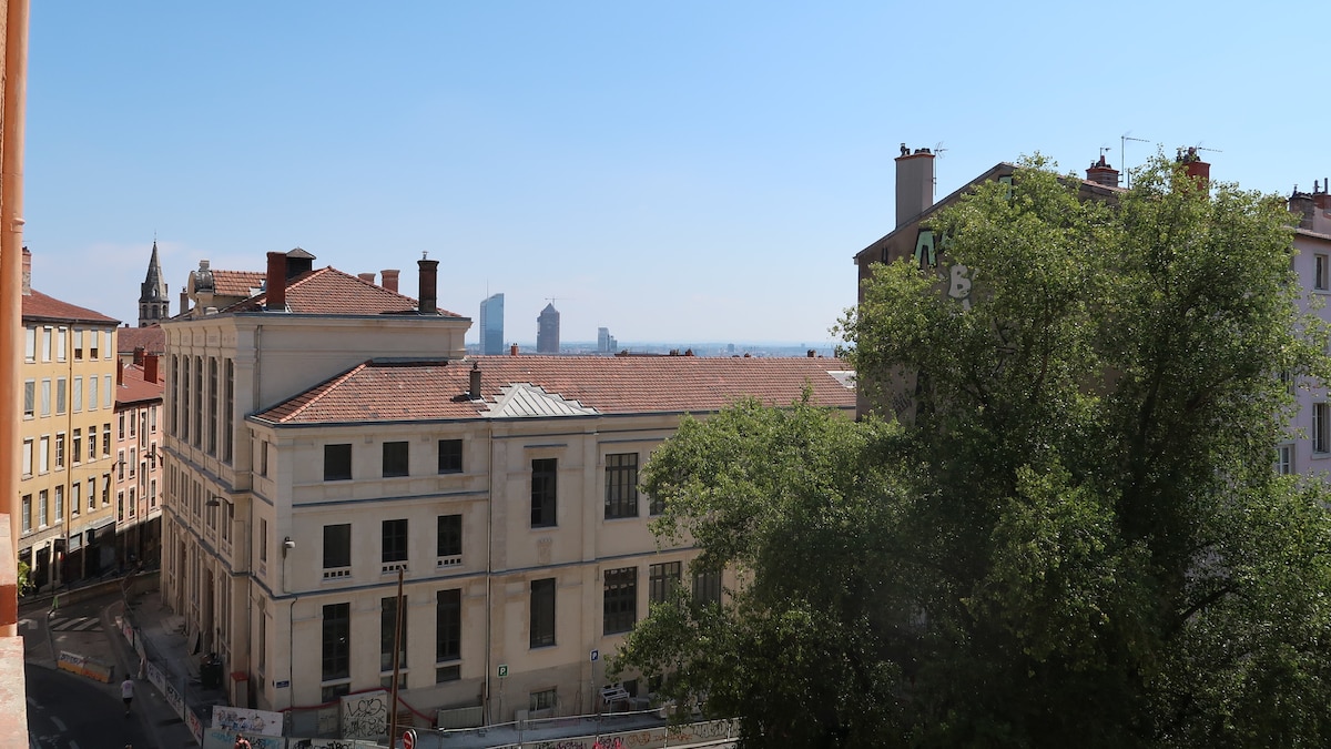 A view from the building showcases a bustling street lined with residential buildings and a mix of urban architecture. In the distance, modern skyscrapers rise against a clear blue sky, and greenery from nearby trees adds a touch of nature to the scene.