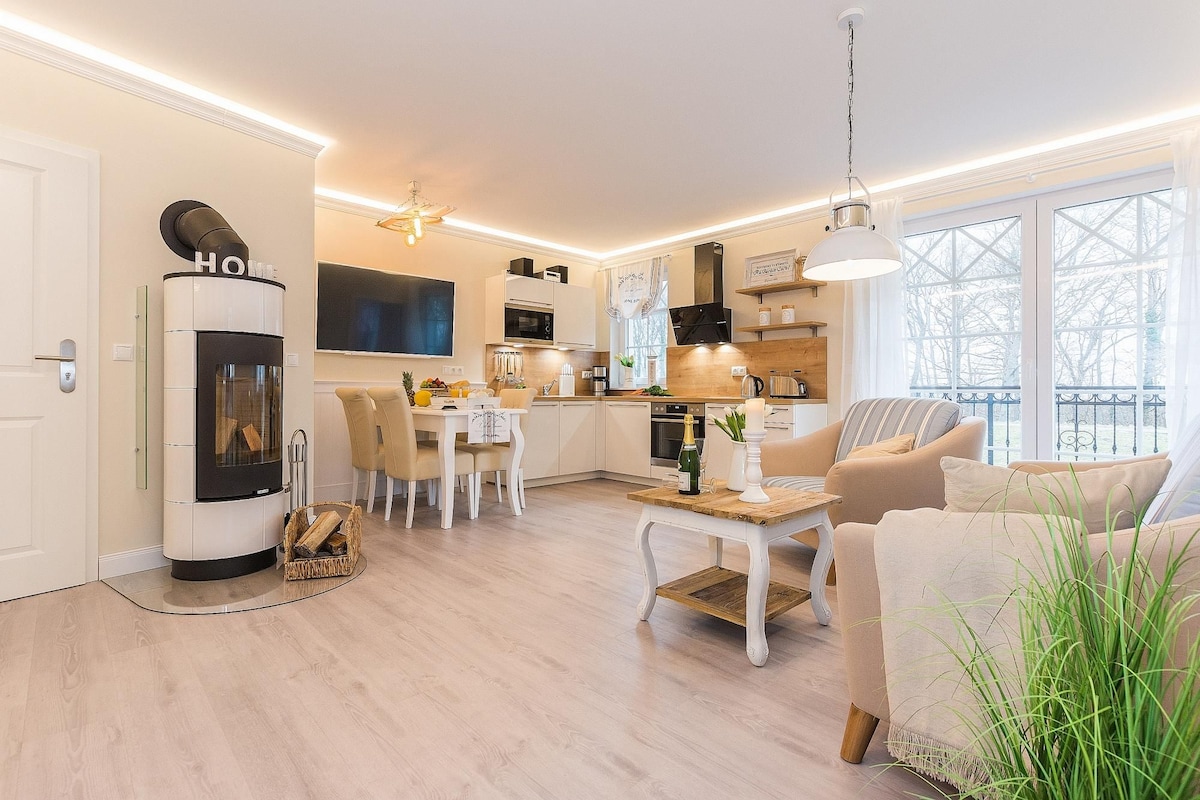 An open-plan living area is highlighted by a modern kitchen with sleek appliances, a dining table, and comfortable seating. A contemporary fireplace adds warmth. Large windows provide natural light and soft views of the surroundings, enhancing the relaxed ambiance of the space.