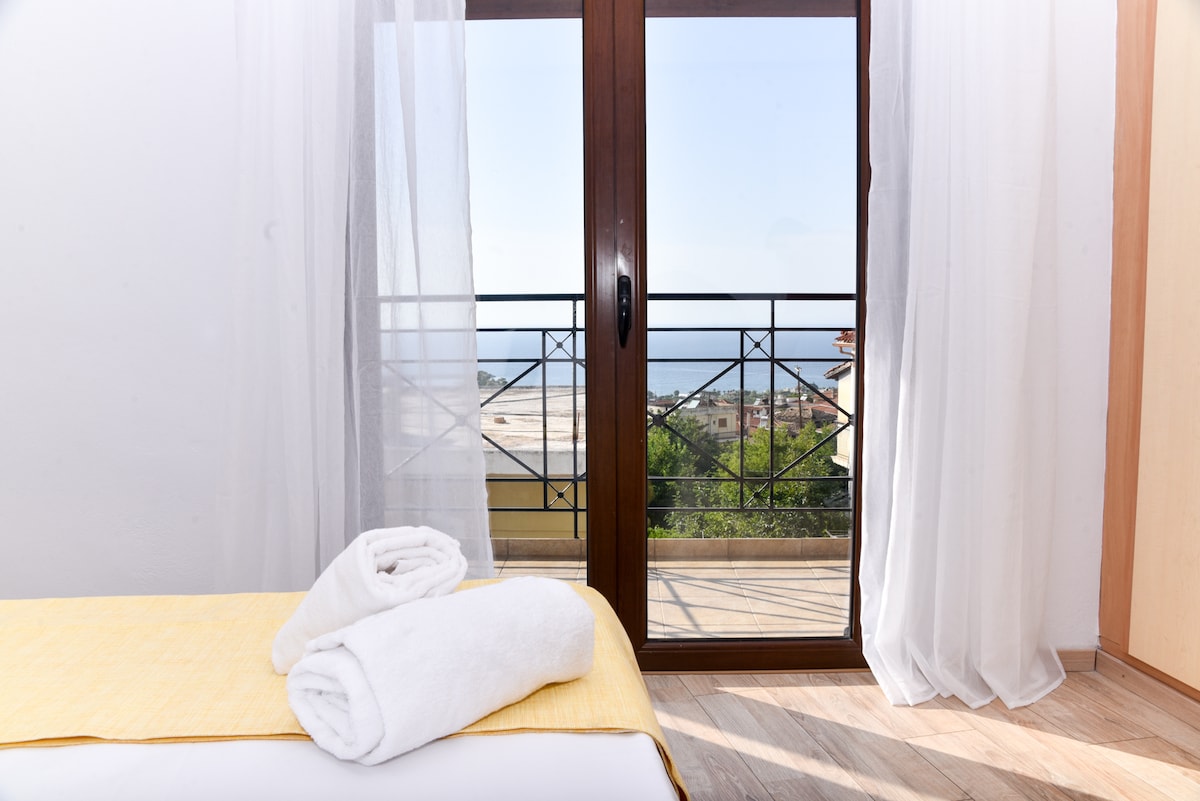 Natural light fills the room through large glass doors, which open to a balcony with views of the Ionian Sea. Two rolled towels are neatly placed on a light-colored bedspread, enhancing the serene atmosphere of the space.