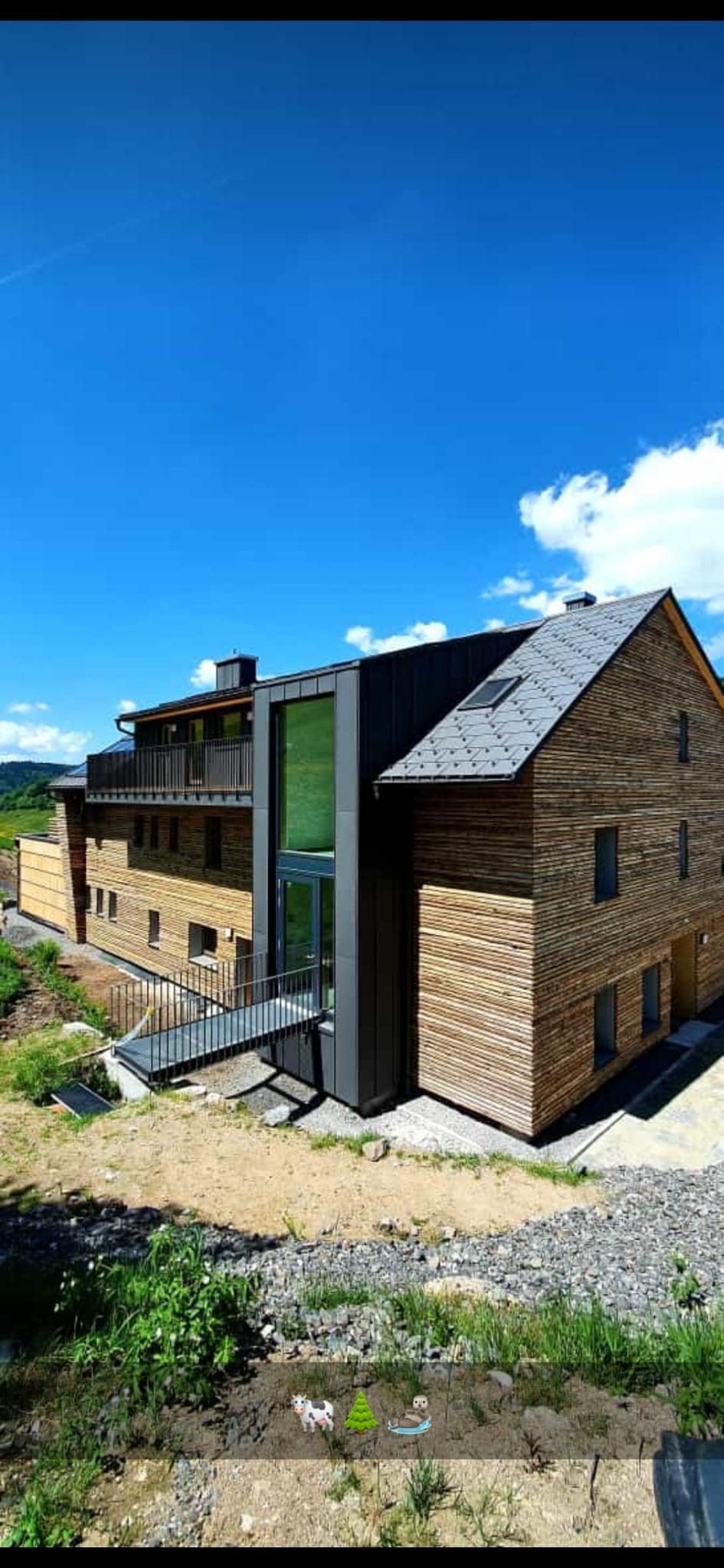 A modern building constructed with natural wood elements is showcased against a clear blue sky. Large windows allow natural light to illuminate the interior. A pathway leads to the entrance, complemented by surrounding greenery and a stone pathway.