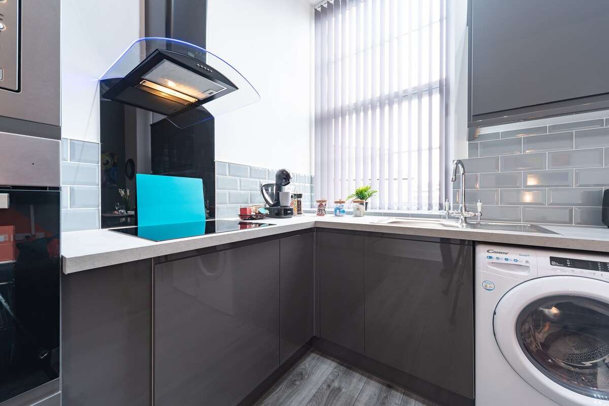 A modern kitchen area features sleek dark cabinets and a stylish countertop. A washing machine is positioned underneath the counter, with a large window allowing natural light. The space showcases a variety of appliances, including a coffee maker and a well-lit sink area.