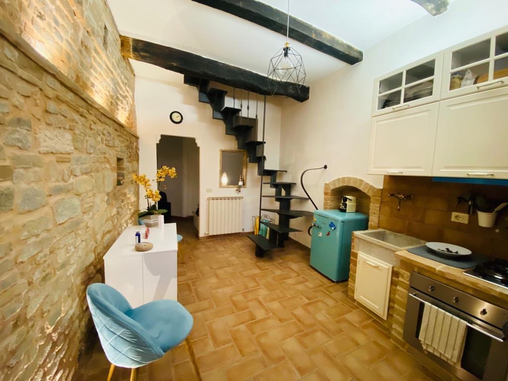 A stylish kitchen area features wooden beams overhead and a spiral staircase leading to an upper level. A bright blue refrigerator stands alongside modern cabinetry and a stovetop. Textured stone walls add character to the cozy space, enhanced by soft lighting.