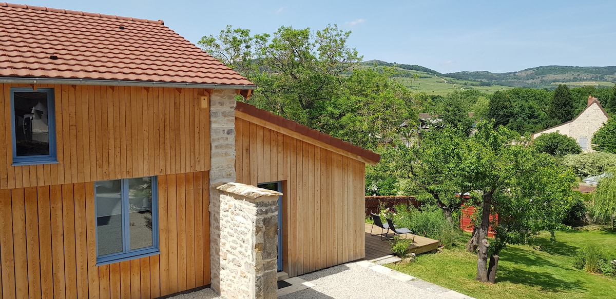The exterior of a contemporary wooden building is highlighted, featuring a sloped roof and large windows. A stone wall partially encloses the entrance. Surrounding greenery includes trees and a landscaped garden, with gentle hills visible in the background under a clear blue sky.