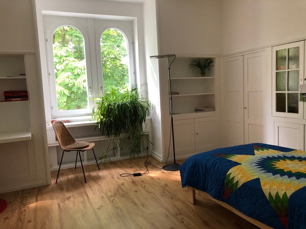 A bright and minimalistic bedroom features a spacious bed adorned with a colorful quilt. Large windows allow natural light to illuminate the room, revealing green plants and a wooden floor. A simple desk and a chair are present, along with shelving for storage.