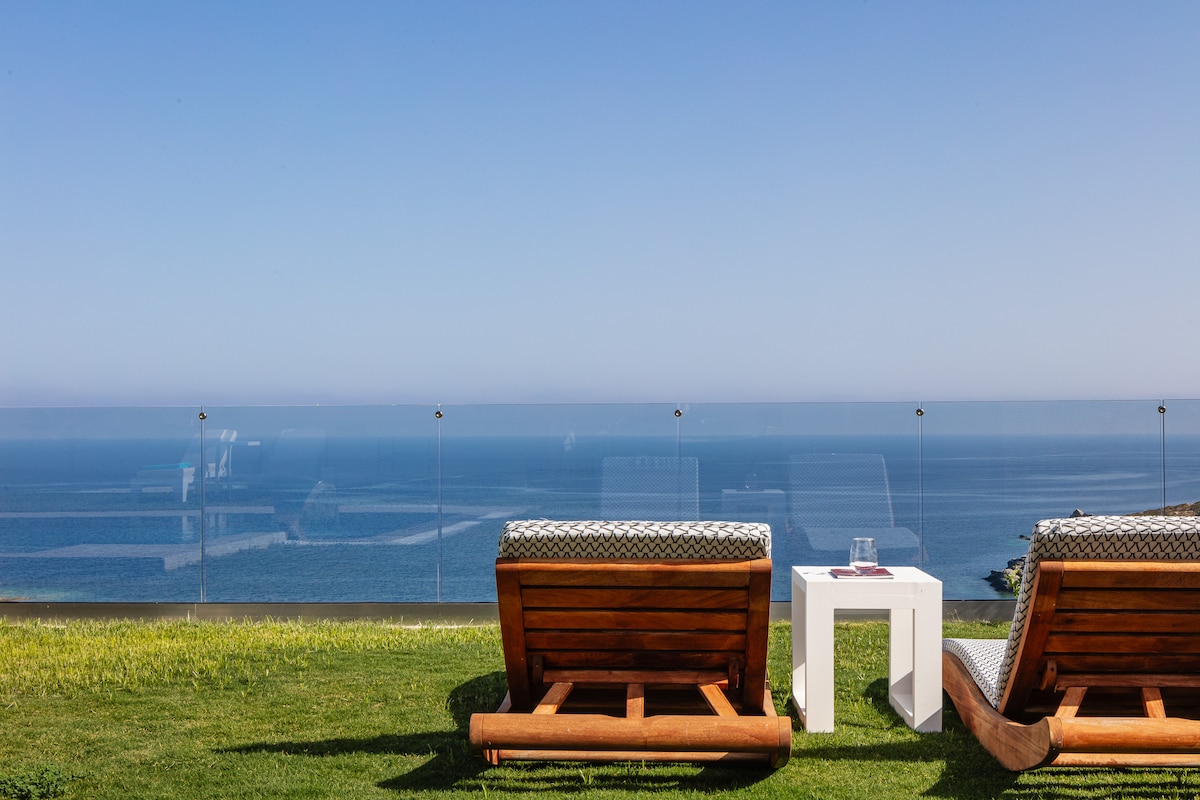 Two wooden loungers are positioned on a manicured lawn, overlooking a serene sea view. A small white table is placed between them, providing a space to enjoy drinks while taking in the unobstructed panoramic vista of the ocean and blue sky.