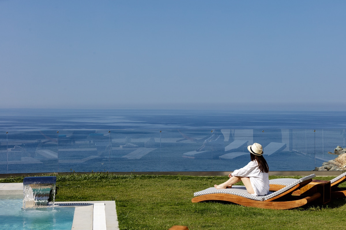 A wooden lounger positioned on manicured grass overlooks a stunning sea view. A gentle waterfall features in the foreground, while the calm waters extend toward the horizon under a clear blue sky.