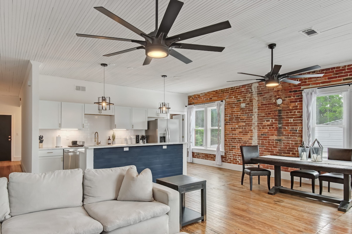 An inviting living space is complemented by two large ceiling fans and exposed brick walls. A comfortable sofa and a dining table for six are present, with natural light filtering through the large windows adorned with light curtains. The kitchen area is streamlined and modern.