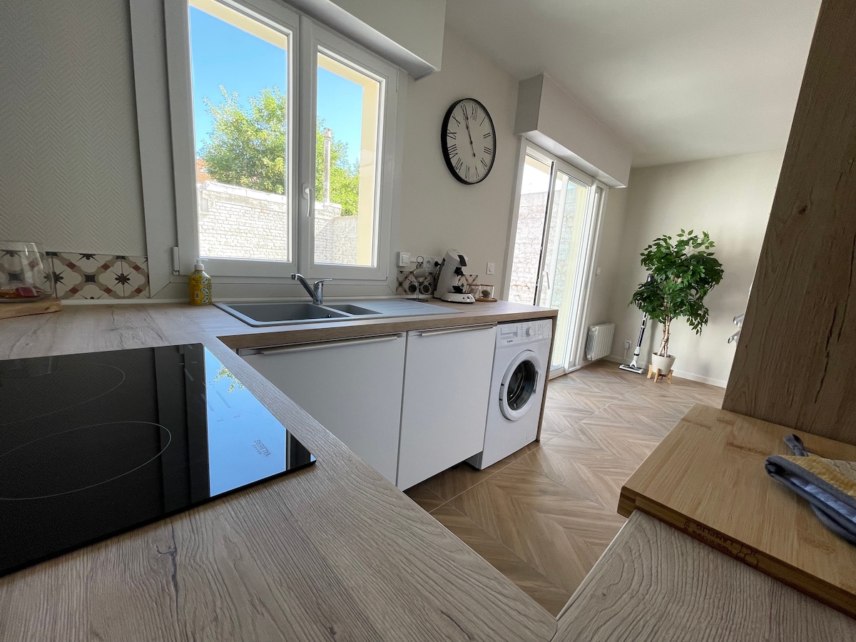 A modern kitchen features a minimalist design with light wood cabinetry and an induction cooktop. A washing machine is integrated into the cabinetry. Large windows allow natural light to illuminate the space, while an indoor plant adds a touch of greenery.
