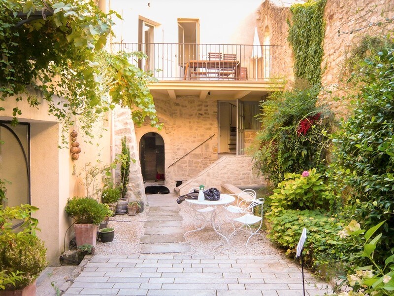 A charming inner courtyard is surrounded by stone walls, displaying lush greenery and various plants. A set of white metal chairs and a small table is visible, while stairs lead to a cozy balcony with wooden seating. The serene space invites relaxation and connection with nature.