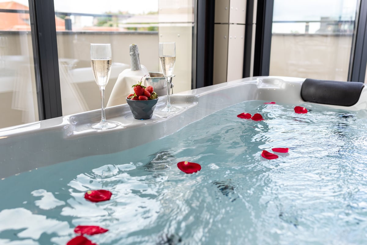 An indoor Jacuzzi is filled with clear water, adorned with floating rose petals. Two glasses of sparkling wine and a bowl of strawberries are positioned on the edge, creating an inviting atmosphere. Large windows provide a view of the surroundings.