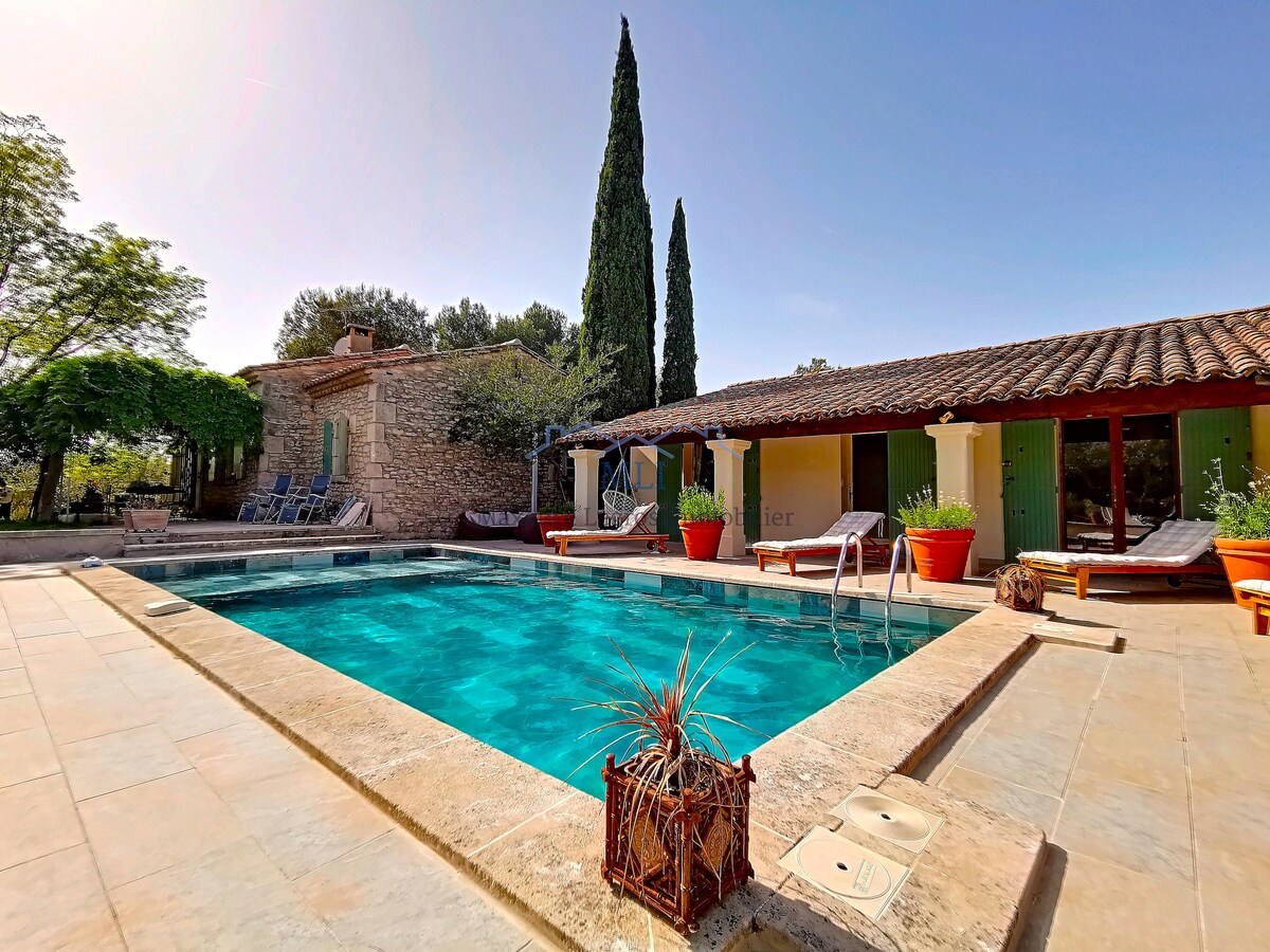 A spacious swimming pool is framed by elegant stone tiles and surrounded by potted plants. A pool house is visible in the background, situated among lush greenery. Lounge chairs are arranged along the poolside, creating an inviting setting for relaxation and leisure.