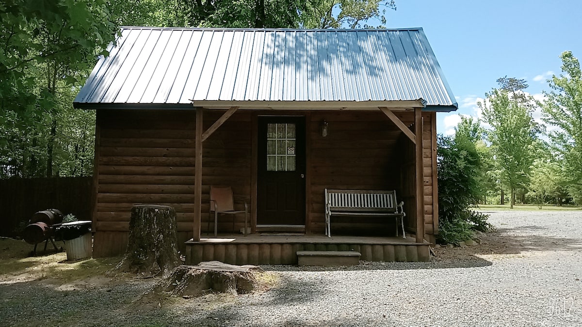 A rustic log cabin with a metal roof features a porch supported by wooden beams. A bench and chair offer outdoor seating in front of the entrance. Surrounding greenery and gravel pathways create a natural setting.