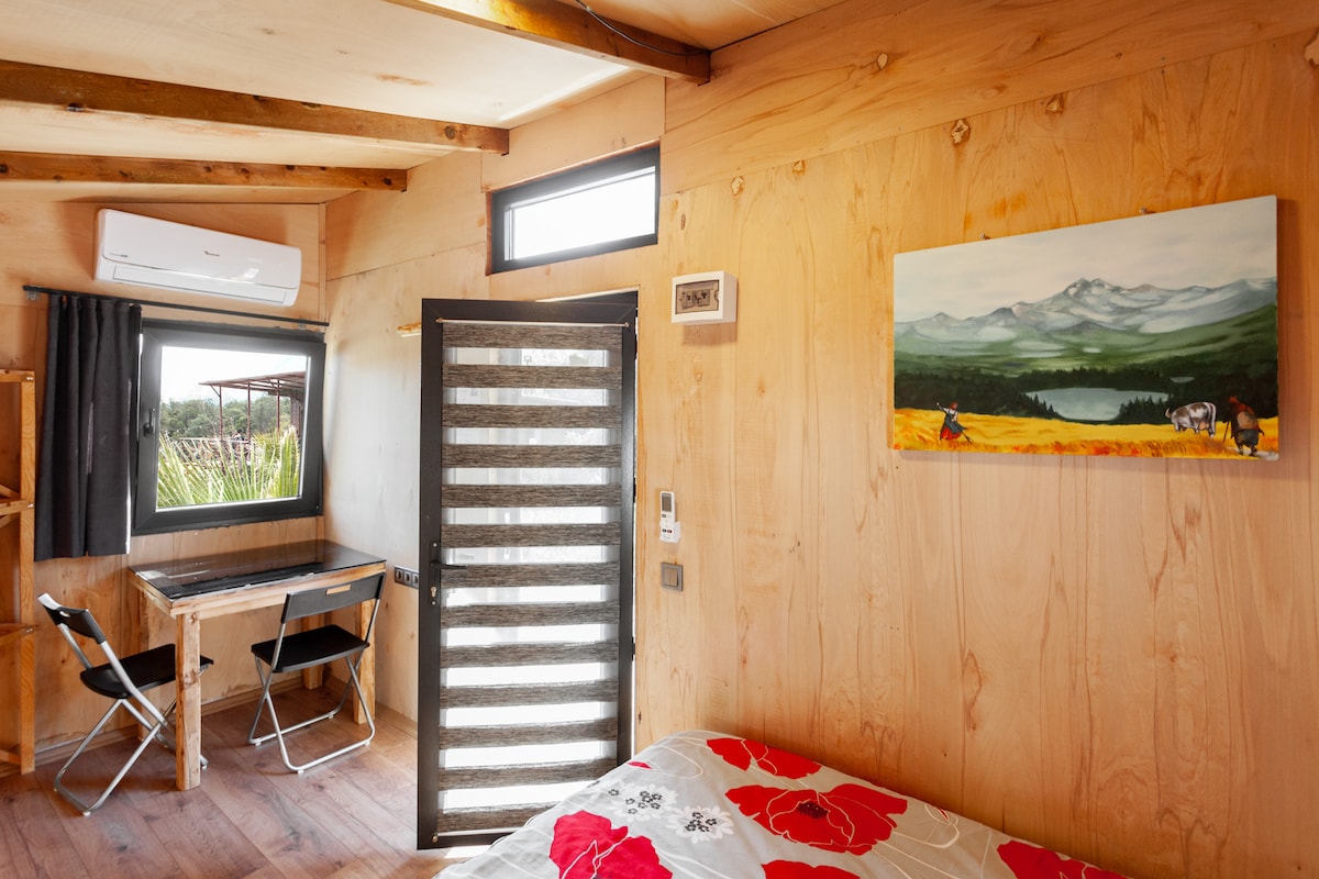 The interior of the bungalow features wooden walls, providing a cozy ambiance. A spacious bed with floral bedding is visible, accompanied by a desk and two chairs. A large window allows natural light, while air conditioning ensures comfort. A scenic painting adds a touch of artistry.