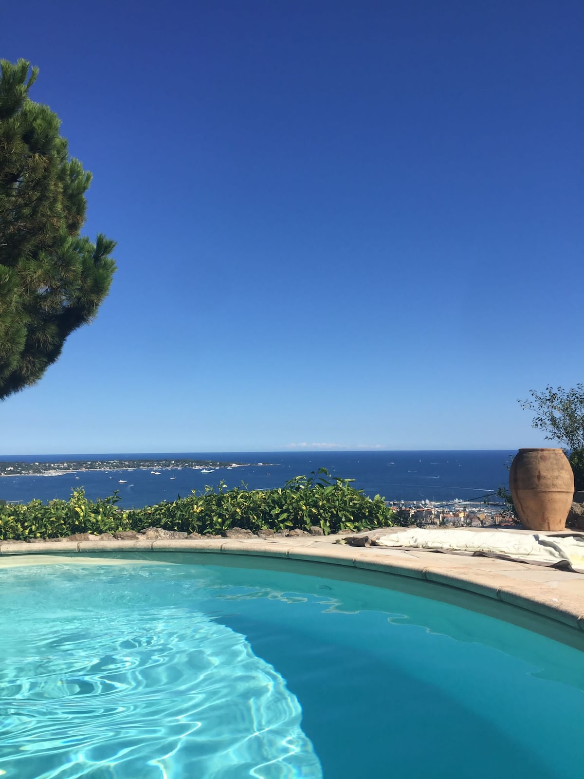 A tranquil pool glistens in the sunlight, surrounded by a stone patio. Lush greenery frames the area, and views of the distant coastline and azure sea stretch beyond. The clear sky enhances the serene atmosphere, providing an inviting backdrop for relaxation.
