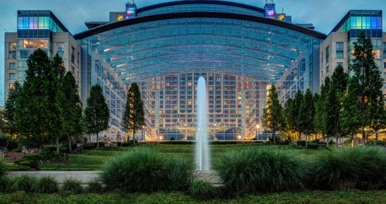 A large modern building with a curved glass structure dominates the scene. Lush greenery surrounds a central fountain, which jets water upward. Illuminated windows reflect the evening light, creating a vibrant atmosphere while tall trees frame the entrance.