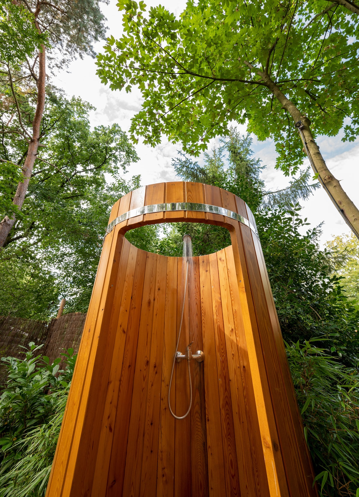 An outdoor shower constructed from natural wood stands tall among lush green trees. The shower features a sleek metal head positioned overhead, with water cascading down, surrounded by vibrant foliage that enhances the natural setting.