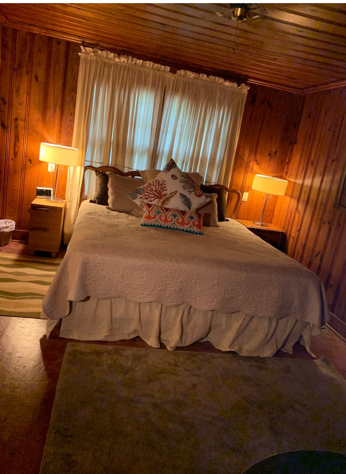 A cozy bedroom features warm wooden paneling and a spacious bed with a light-colored quilt. Two bedside lamps provide soft light, and a decorative pillow adds a touch of color. Light streams in through the flowing curtains, enhancing the inviting atmosphere.