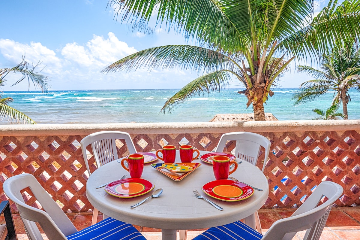 An outdoor dining area is set with a round table featuring colorful plates and cups. Nearby palm trees frame the view of the Caribbean Sea, while waves gently lap at the shore, creating a serene backdrop for meals.