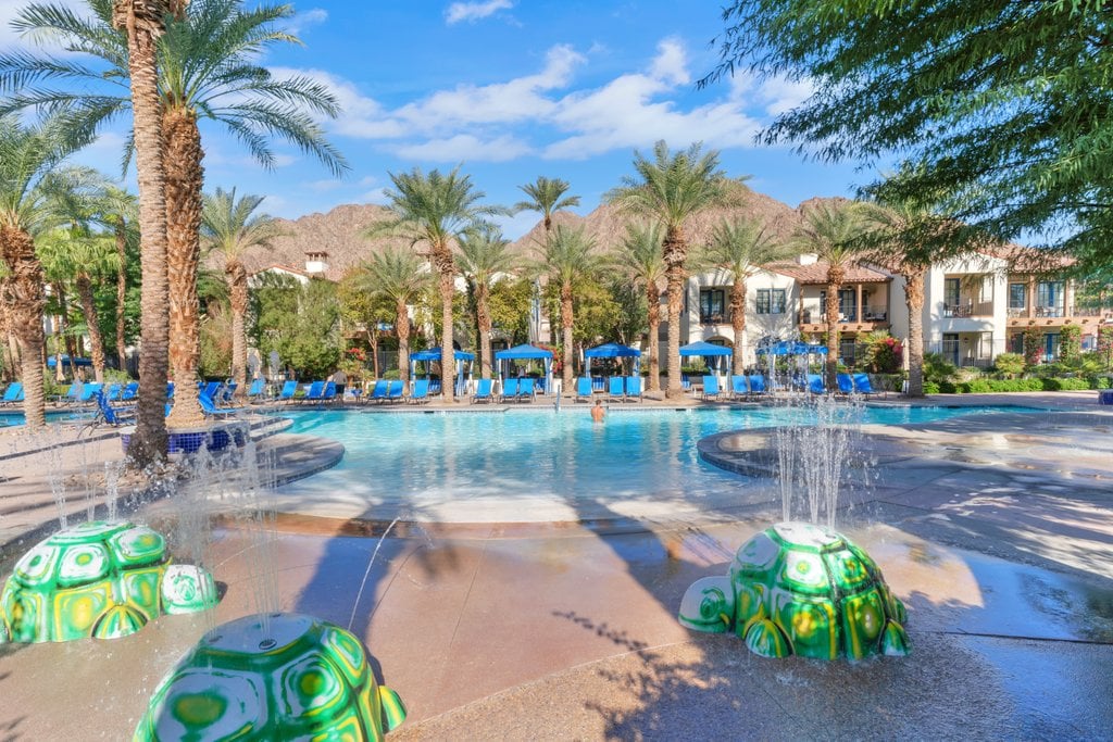 A serene pool area is surrounded by tall palm trees, with cozy blue lounge chairs positioned underneath shaded umbrellas. Decorative green turtle sculptures sit at the pool's edge, while gentle water features add a calming element to the vibrant landscape in the background.