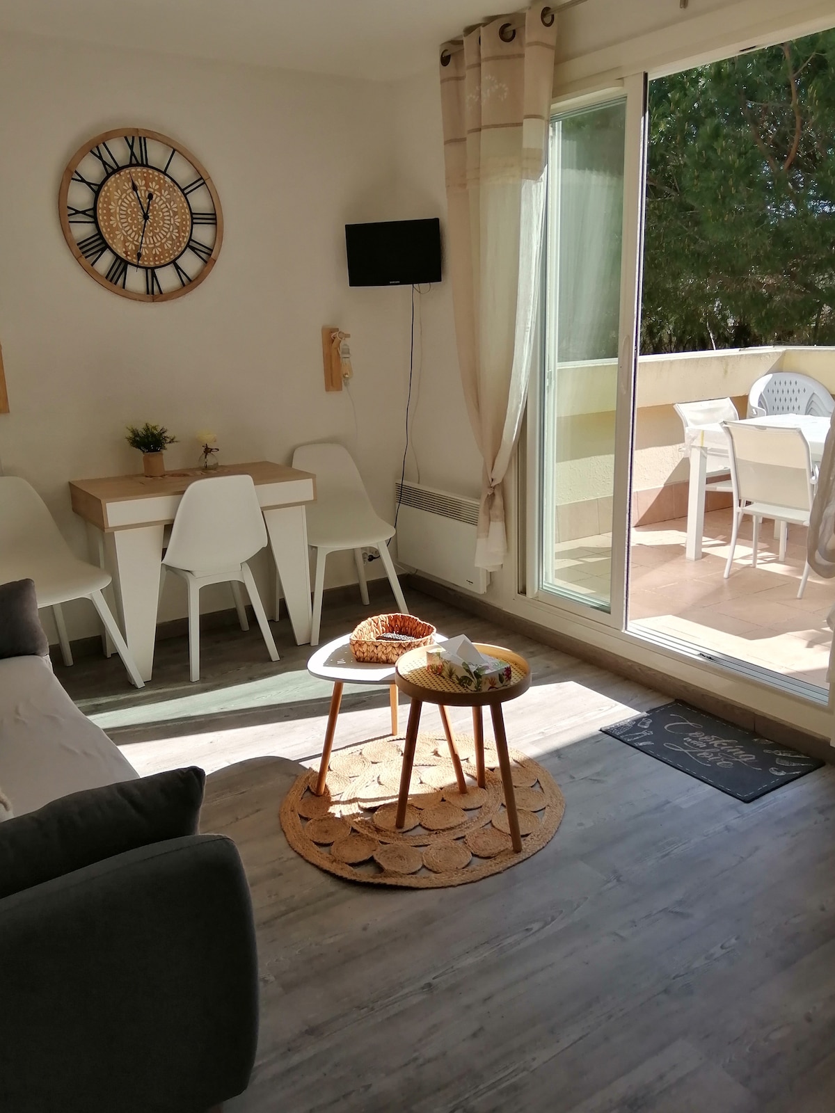 A bright living area features a neutral-toned sofa facing a small round table with snacks. A dining area with white chairs is present, adjacent to a glass sliding door leading to a balcony. Natural light illuminates the space, complemented by a wall clock.