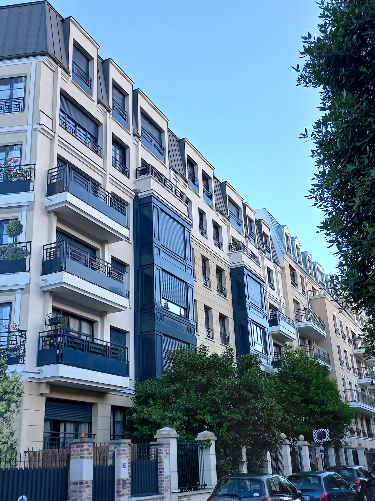 The modern building features multiple stories with a combination of light and dark materials. Balconies with railings are visible on several floors, adorned with potted plants. Lush greenery is present at the base, offering a natural contrast to the architectural design.