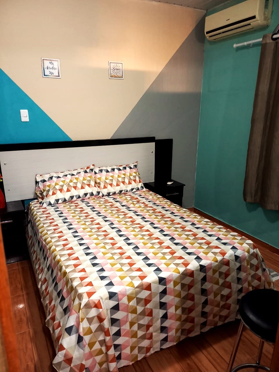 A compact bedroom is presented, featuring a neatly made bed adorned with a geometric pattern in warm colors. The walls are painted in soothing teal and neutral tones, and a small nightstand is positioned beside the bed. Natural light filters through a partially draped window.