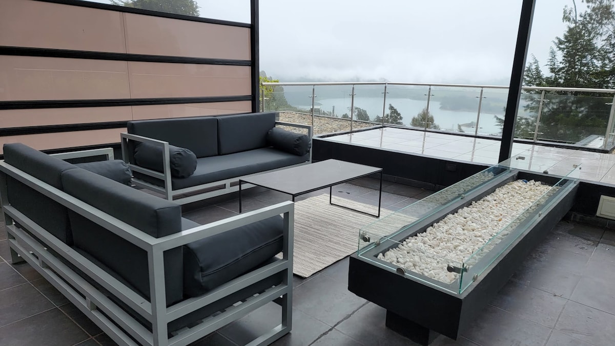 A modern seating area is presented with a sleek black sofa set arranged around a minimalist coffee table. A long fire pit with white stones serves as a centerpiece, while a view of the lake and misty landscape is framed by expansive glass walls.