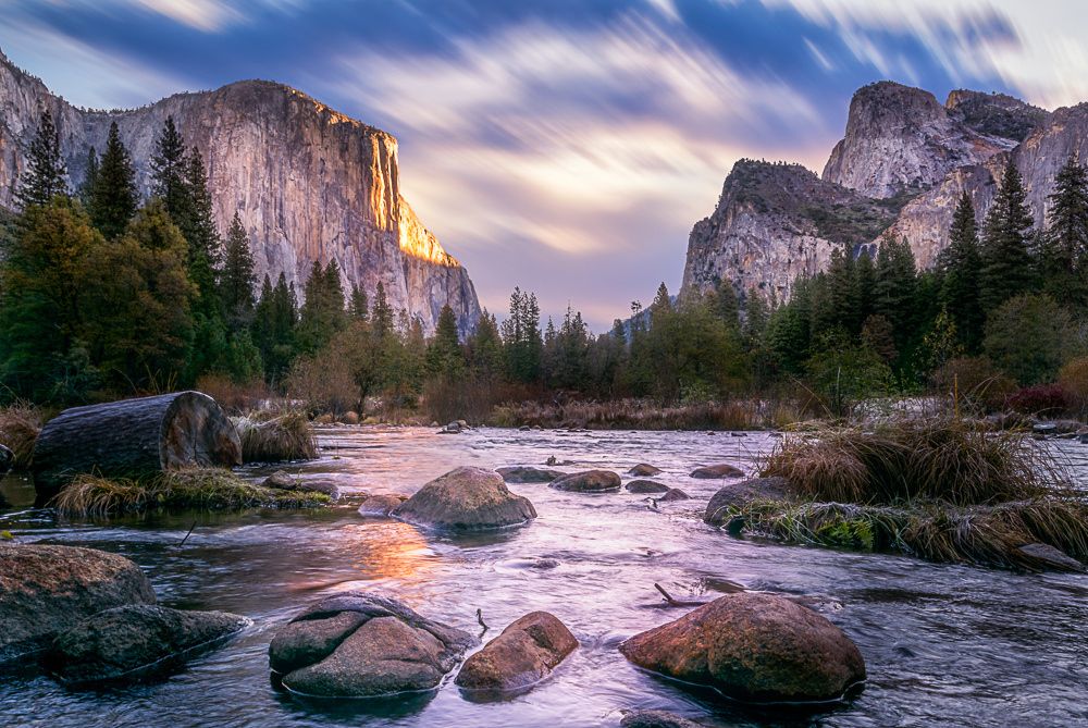 A serene river flows through a valley, surrounded by towering cliffs and lush greenery. Smooth stones are visible beneath the water's surface, while the sky showcases a gradient of colors, indicating the transition of day. Gentle ripples create a tranquil atmosphere.