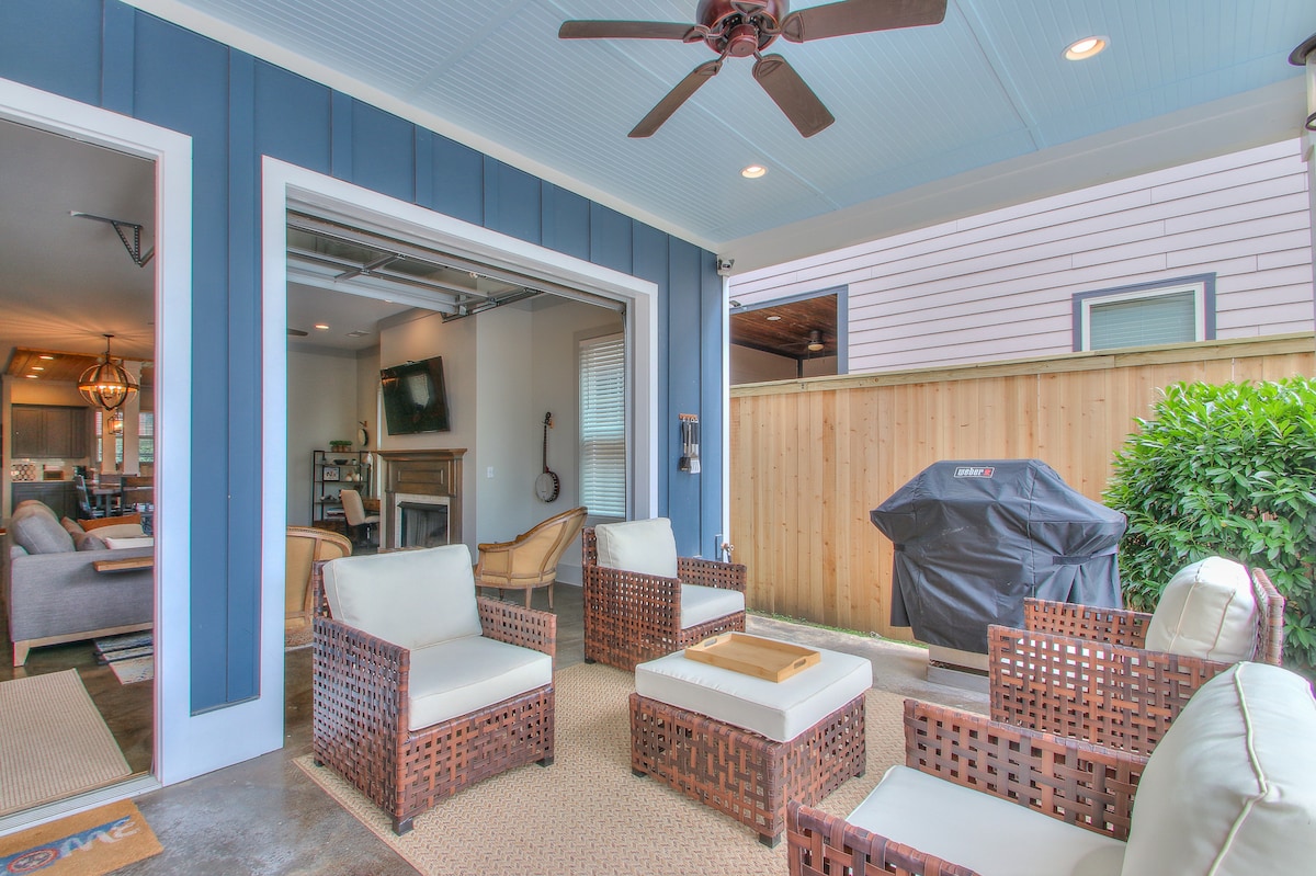 An outdoor covered patio features comfortable seating arranged around a central table. A gas grill is visible in the corner, and a ceiling fan provides airflow. The space connects seamlessly to the indoor living area through a large roll-up door, allowing for versatile entertaining.