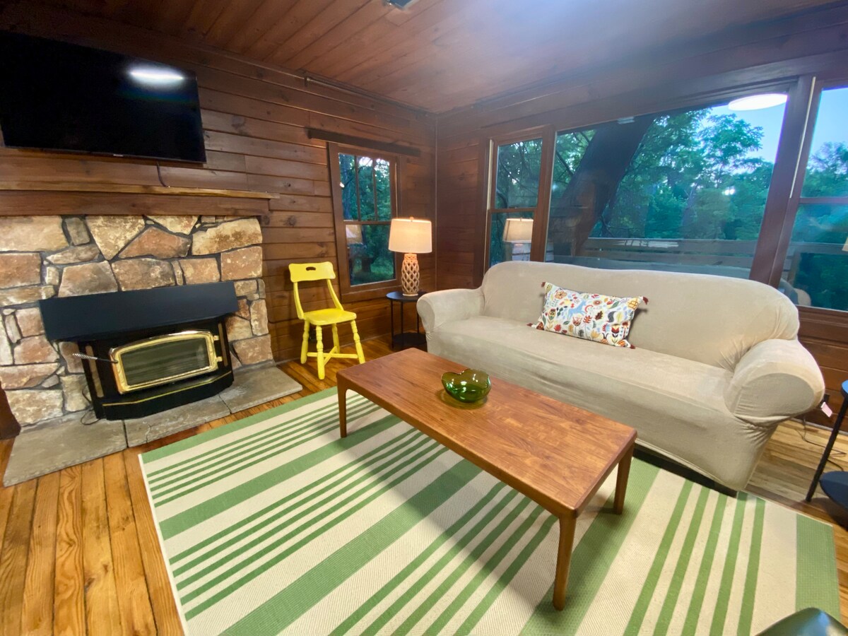 A cozy living area features a stone fireplace and large windows, allowing natural light to fill the space. A comfortable sofa is complemented by a wooden coffee table. A yellow chair and a lamp add a touch of warmth to the rustic wooden interior.