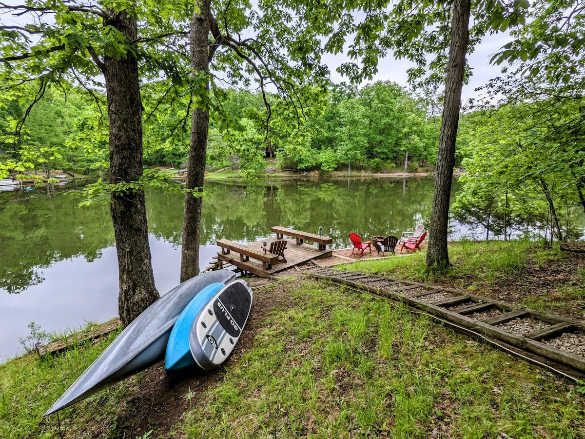 A serene lake view is presented, with a canoe and paddleboard resting on the grass. Wooden benches and red adirondack chairs are arranged along the water's edge, surrounded by lush green trees and reflecting the calming atmosphere.