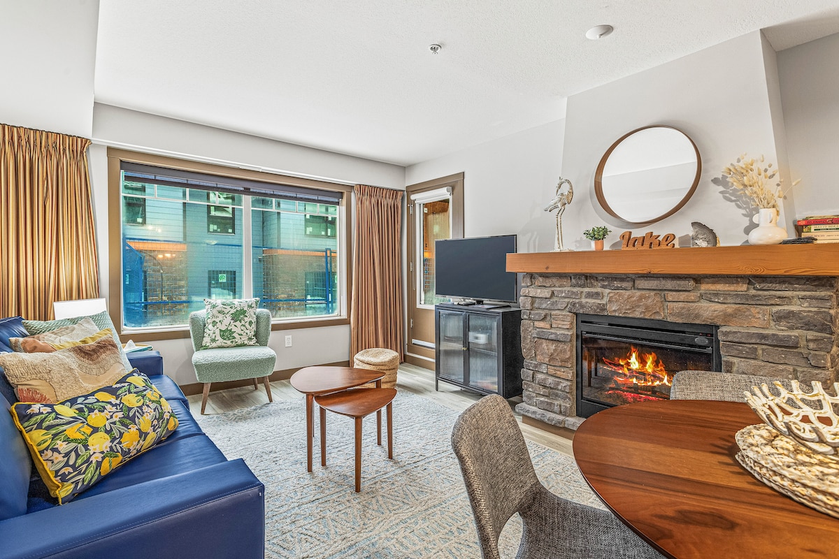The inviting living area features a blue sofa paired with a patterned lounge chair. A rustic stone fireplace is set beneath a television. Natural light floods the space through large windows dressed in warm curtains, offering views of the outdoor scenery.