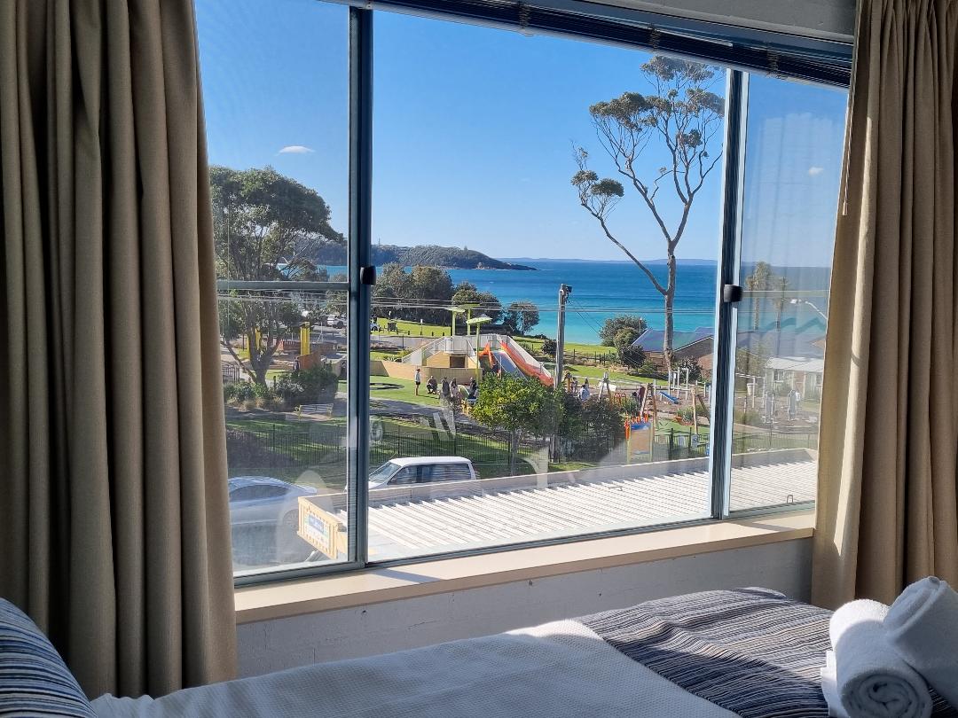 A bright view is captured from the window, showcasing ocean vistas complemented by a beachside playground. The scene features green trees and calm waters under a clear sky, with light filtering through the window curtains and emphasizing the inviting atmosphere of the space.