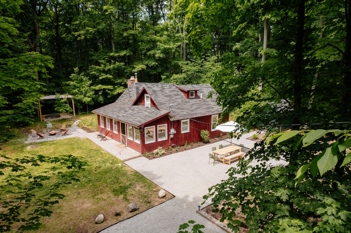 A charming cottage is nestled in a lush green forest, showcasing a red exterior and a sloped roof. An outdoor dining area is visible on a gravel patio, with seating and an umbrella, while a fire pit area is situated nearby among the trees.